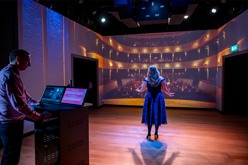 A female student performing in front of a fake theatre audience in a simulator, with a man managing a tablet and screens in the background.